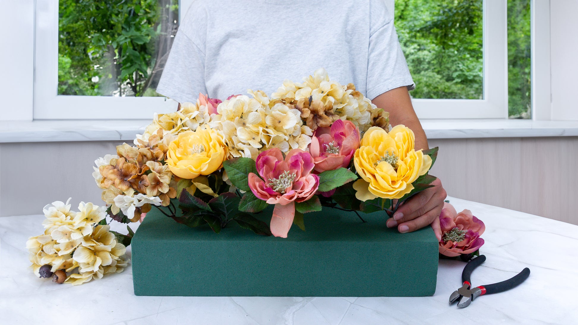 Person arranging a colorful floral centerpiece with peonies and roses on a green floral foam base.