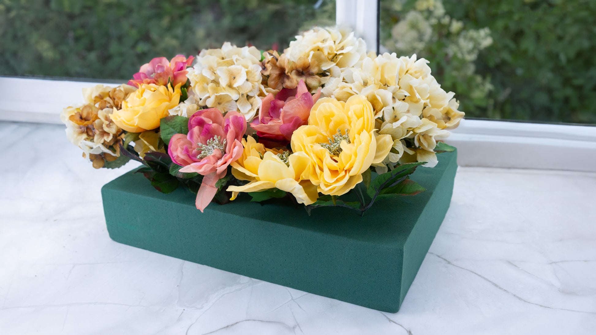 Artificial flower arrangement with yellow, pink, and cream blooms in green floral foam.