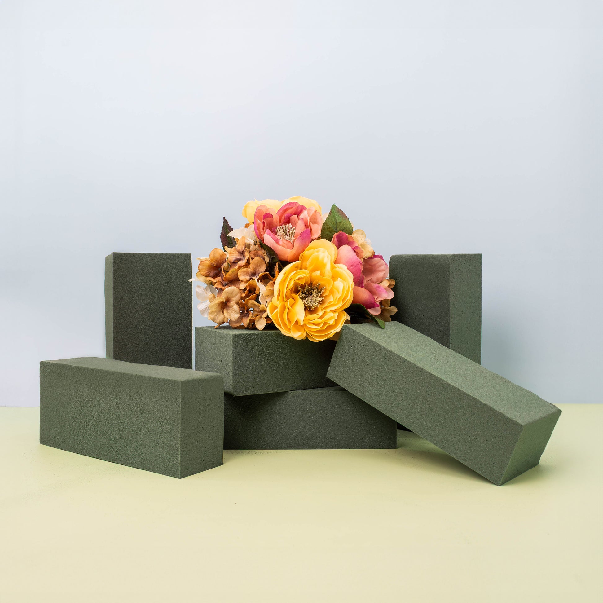 A bouquet of vibrant orange and pink flowers resting on green floral foam blocks arranged on a light-colored surface.