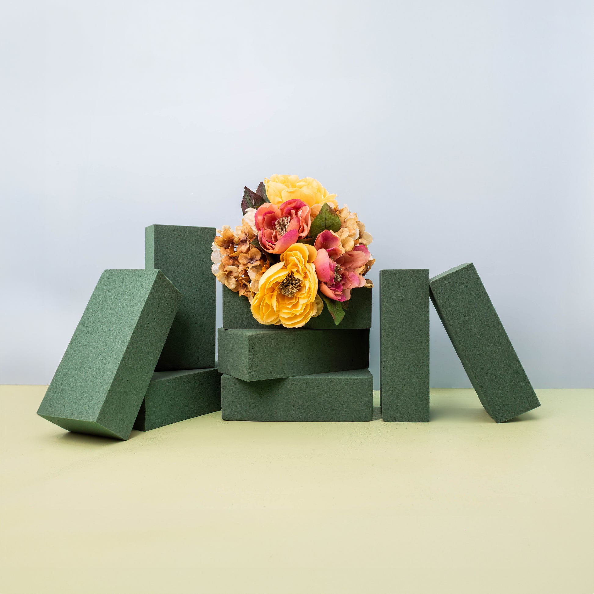A stack of green rectangular floral foam blocks arranged around a small bouquet of orange and pink artificial flowers, all set on a light green surface with a light blue background.