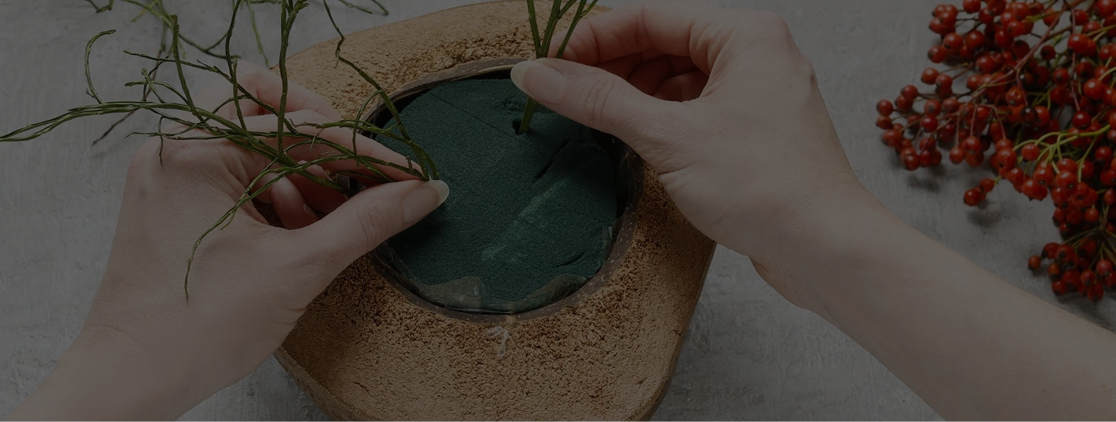 Hands arrange green foliage in a round container with floral foam, creating a display; red berries accent the background
