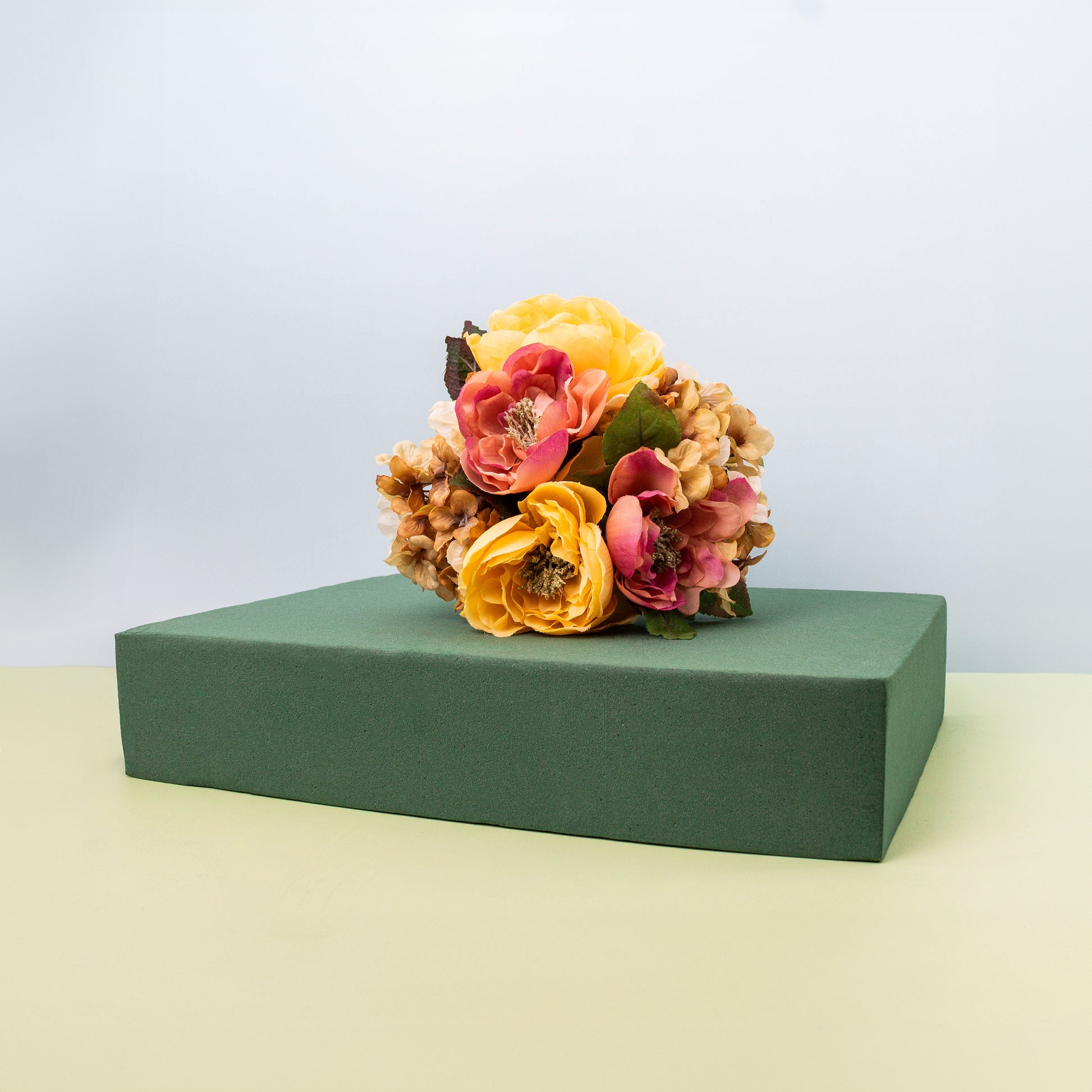 Artificial flower bouquet featuring yellow and pink blooms, possibly peonies or ranunculus, arranged on a rectangular block of green floral foam.