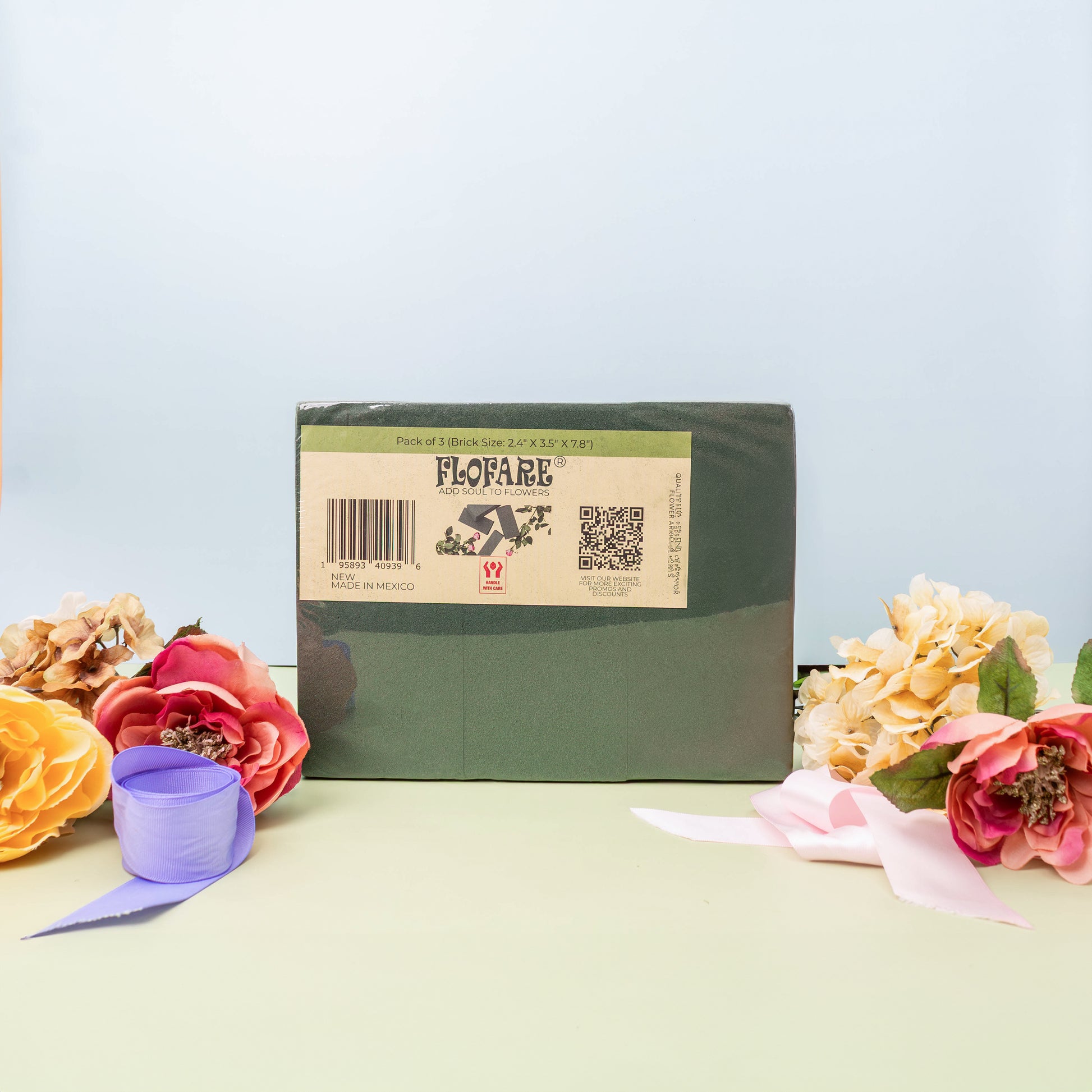 A pack of three green FLOFARE floral foam blocks with a label, surrounded by colorful artificial flowers and ribbons on a light-colored surface.