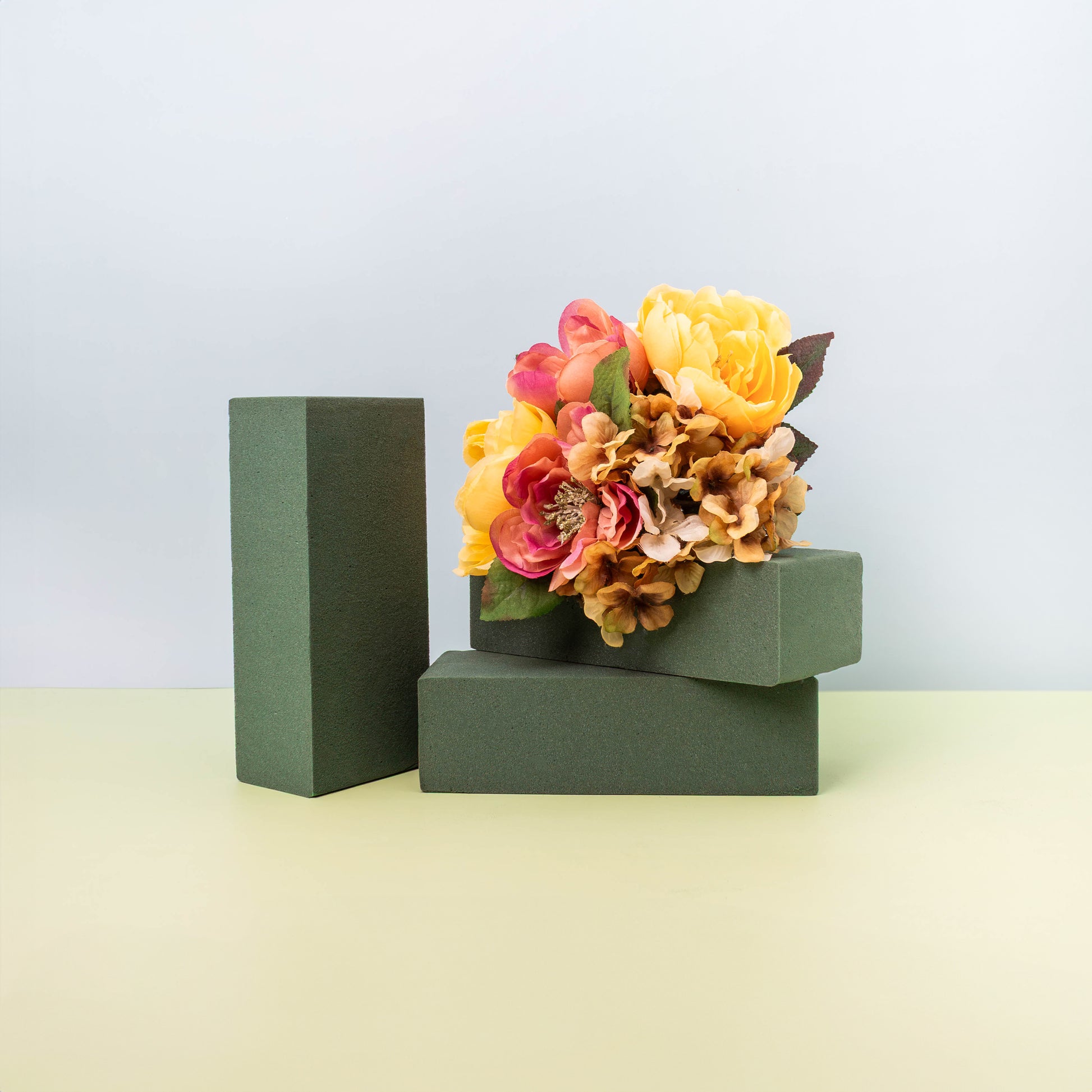 Stack of dark green floral foam blocks, with a vibrant bouquet of orange and pink artificial flowers arranged on the top block.