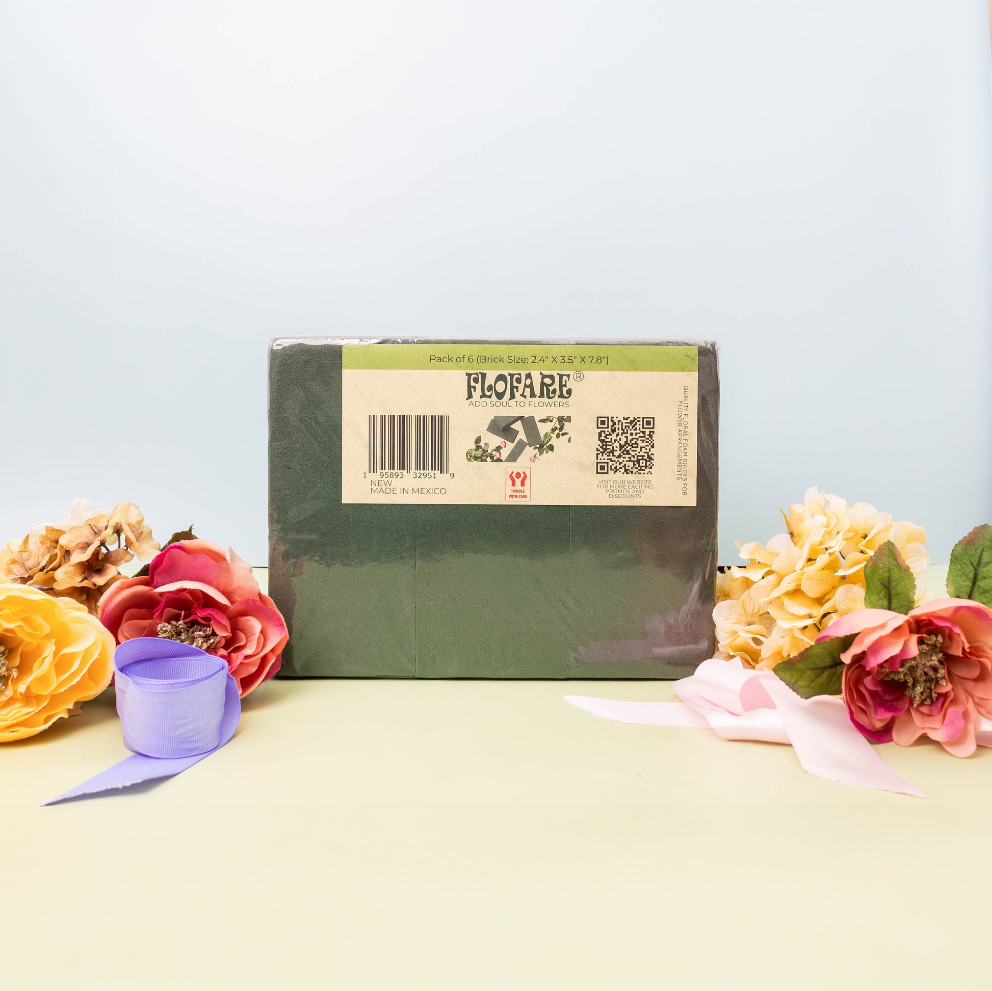 A package of green Flofare floral foam blocks, labeled "FLOFARE," sits on a light green surface, surrounded by colorful artificial flowers and ribbons.