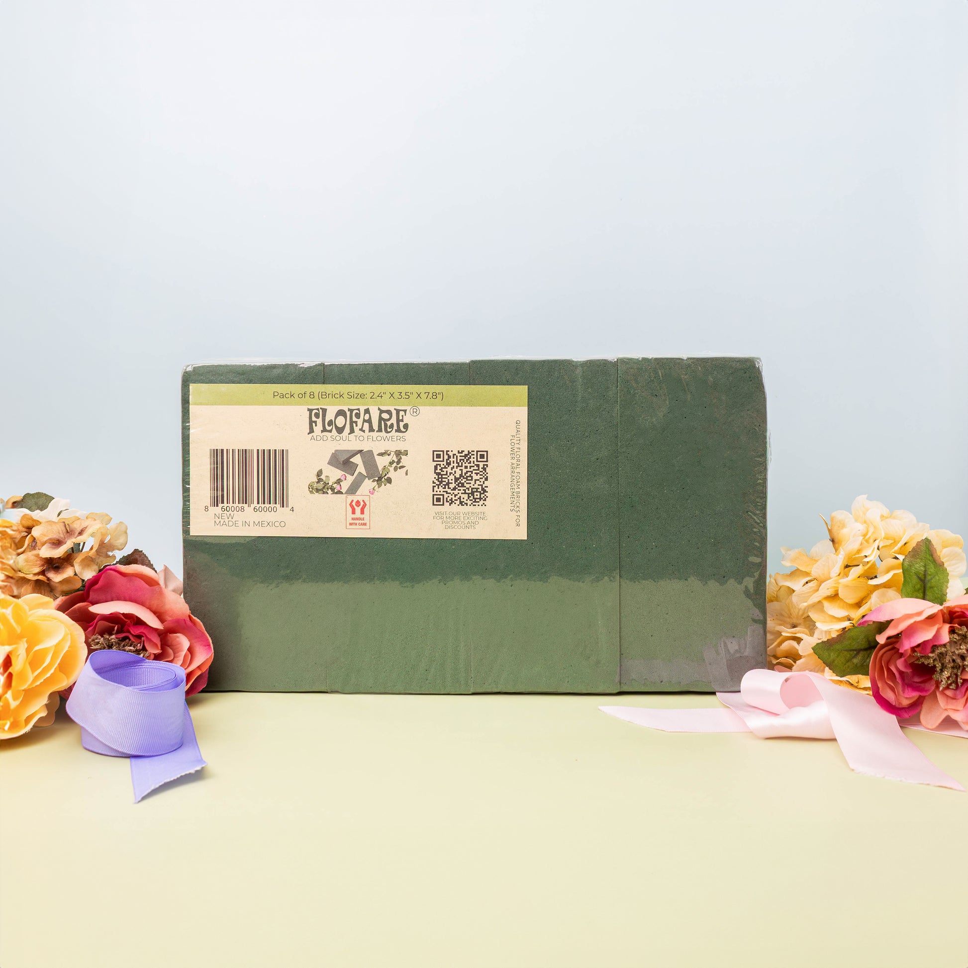 A green FLOFARE floral foam block in its packaging, surrounded by bouquets of pink, yellow, and orange flowers, and light pink and purple ribbons, on a green surface with a light blue background.
