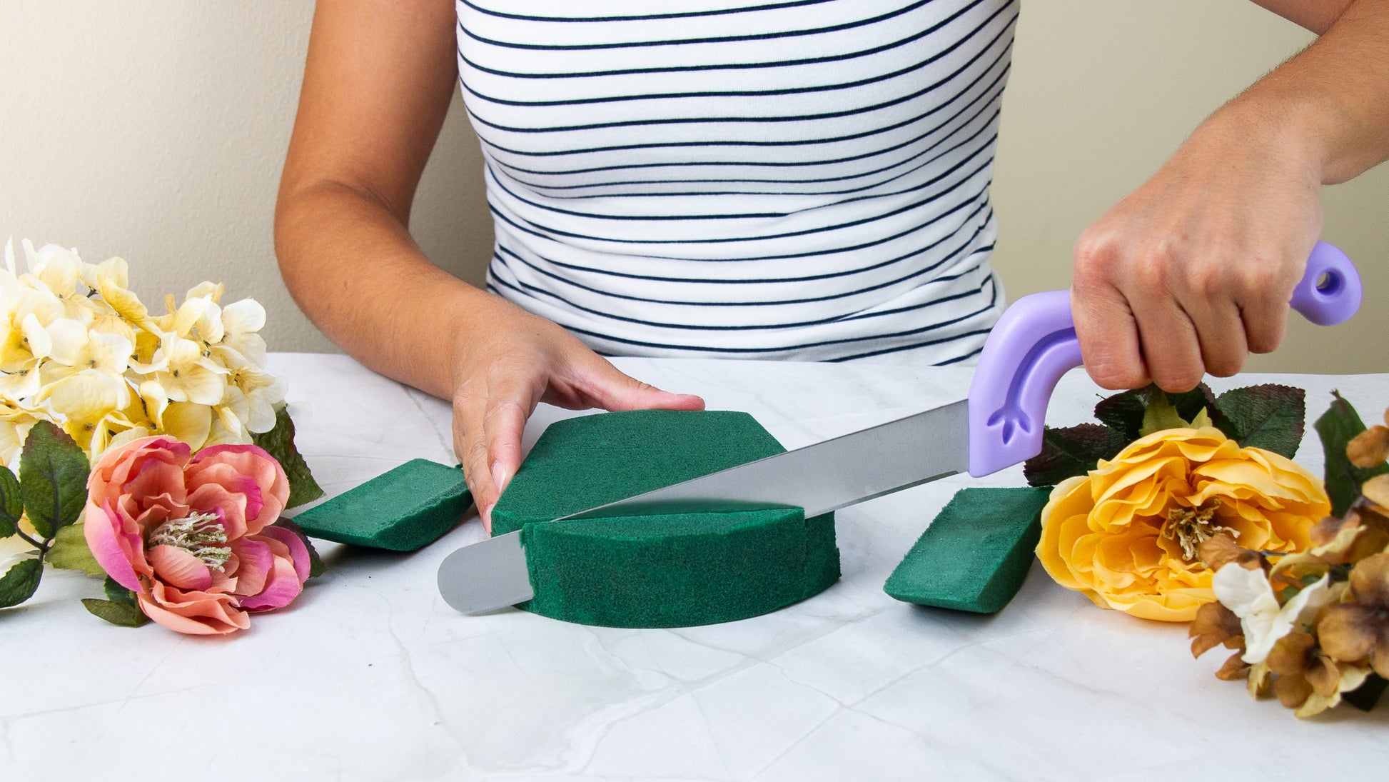 Woman cutting a green floral foam block with a purple-handled knife, preparing it for flower arrangement, with colorful flowers nearby.
