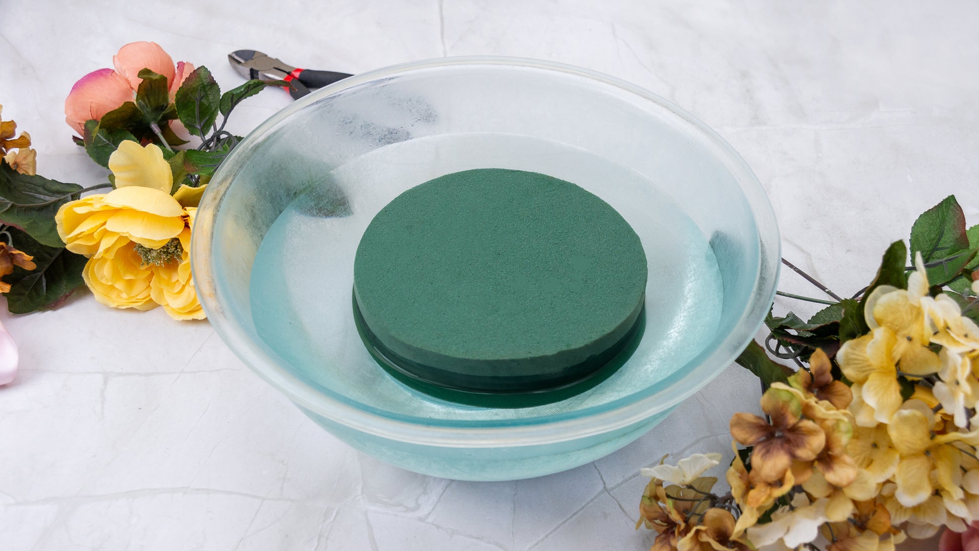 Green round floral foam soaking in a clear bowl of water with various artificial flowers and tools nearby for arrangements