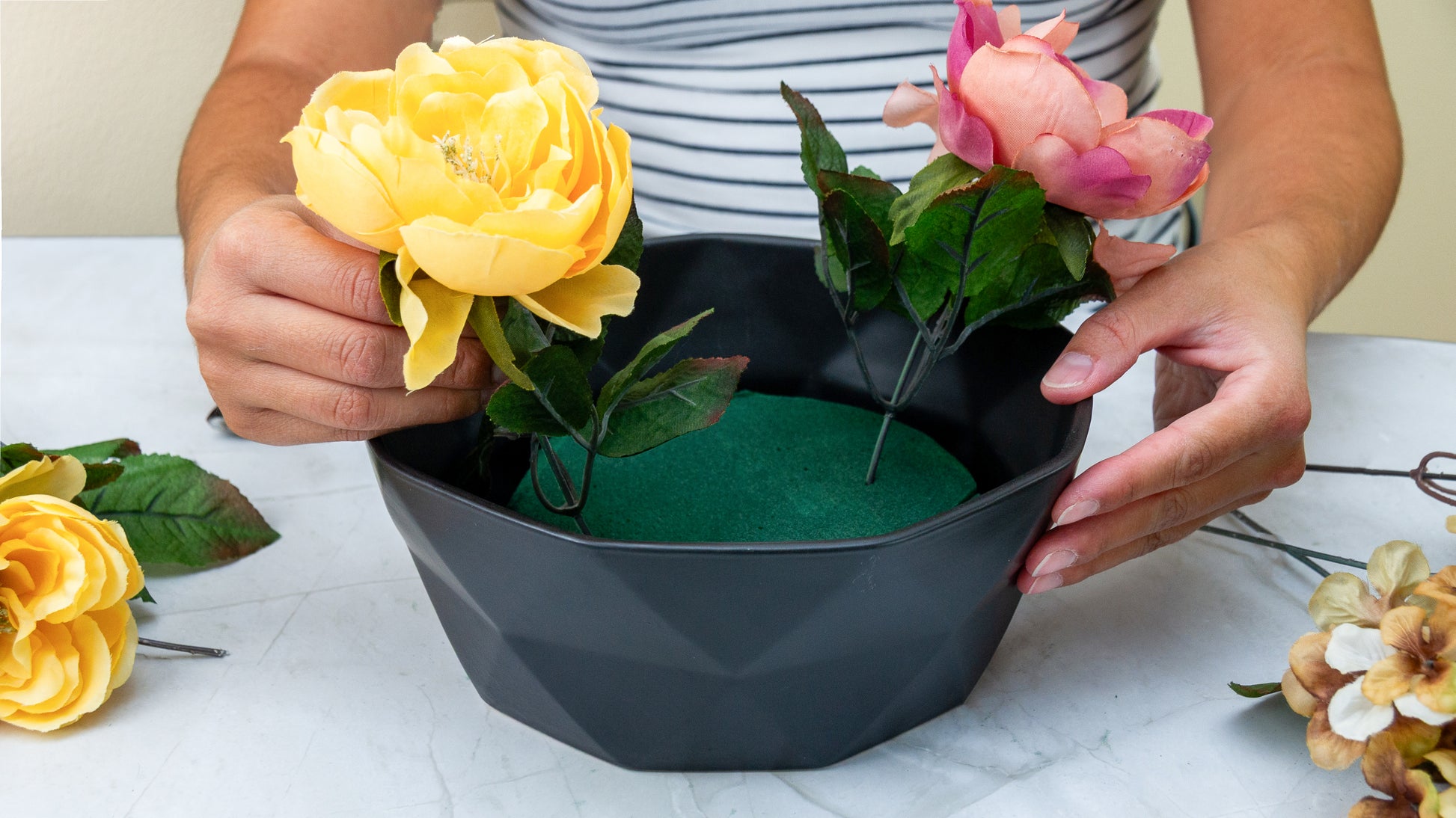 Person arranging artificial yellow and pink flowers into a dark gray geometric planter containing green floral foam