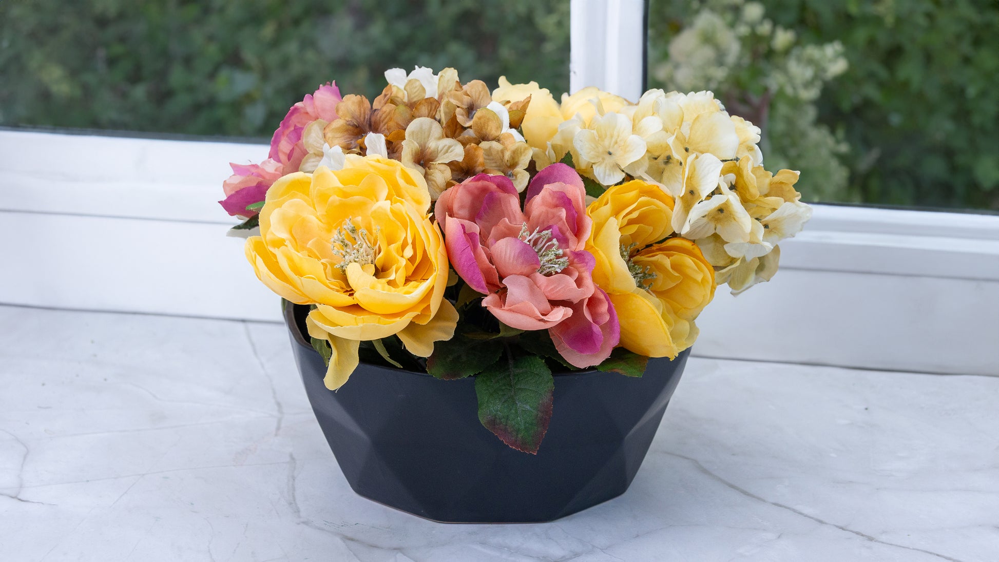 Colorful artificial floral arrangement in a black geometric vase on a white windowsill, against a blurred outdoor background
