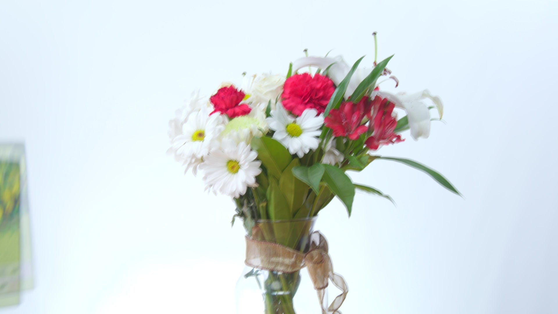 A vibrant mixed flower bouquet with red carnations, white daisies, and lilies in a clear vase with a ribbon
